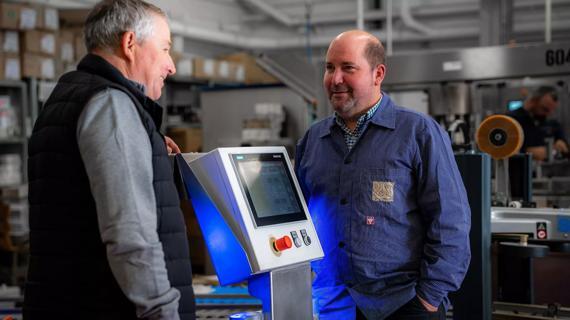 Zwei Männer stehen und unterhalten sich neben einer industriellen Schalttafel in einer Lagerhalle, umgeben von Regalen mit Kisten und Maschinen.