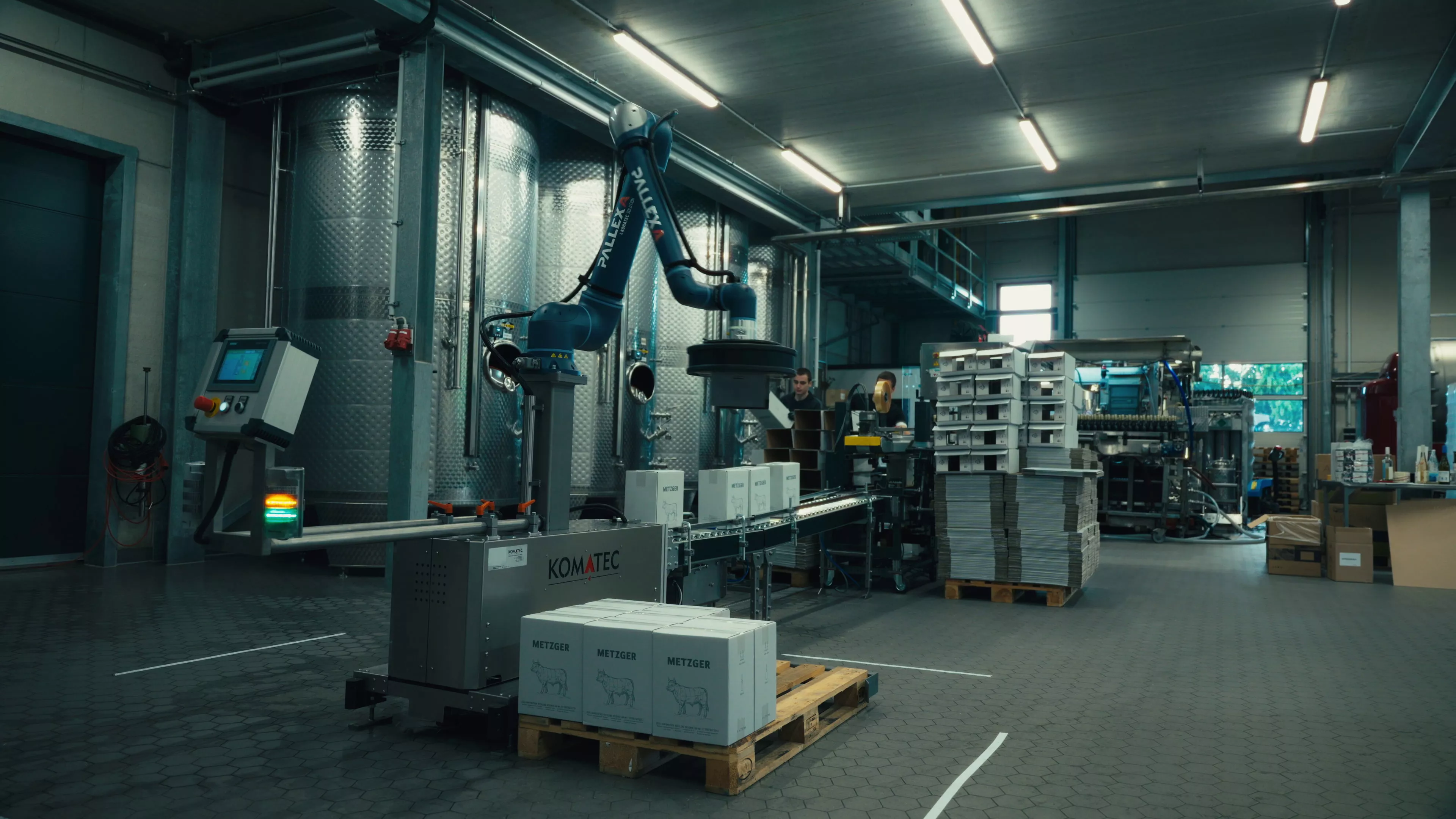 Ein Roboterarm in einer Industrieanlage befördert Kisten auf ein Förderband, das von Metalltanks, gestapelten Tabletts und verschiedenen Geräten umgeben ist.