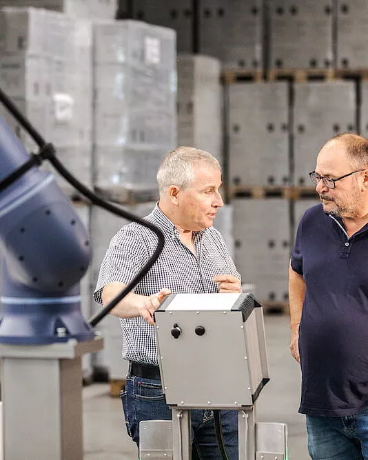 Zwei Männer stehen in einer Lagerhalle in der Nähe einer kompakten Palettieranlage und unterhalten sich. Einer gestikuliert, während sie diskutieren. Im Hintergrund sind Kisten ordentlich gestapelt.