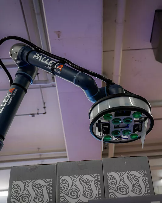 Ein Roboterarm mit der Bezeichnung "PALLEXA" palettiert und hebt eine Kiste mit einem Sauggreifer in einer Industrieumgebung, in der sich unten kompakt gestapelte gemusterte Kisten und oben eine Decke mit freiliegenden Rohren befinden.