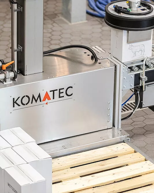 Eine Maschine mit der Aufschrift KOMATEC verarbeitet Kartonagen auf einem Fördersystem. Im Vordergrund sind mehrere weiße Kartons auf einer Holzpalette gestapelt. Die Maschine steht auf einem grauen Fliesenboden.